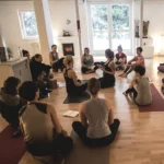 Eine Gruppe von Menschen sitzt auf Yogamatten in einem hellen Raum und hört einer Yogalehrerin zu.
