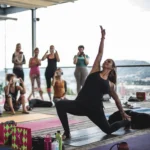 Eine Frau macht Yoga auf einer Terrasse, während mehrere Personen im Hintergrund zuschauen und Fotos machen.