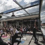Eine Gruppe von Menschen praktiziert Yoga auf einer Terrasse unter einem Glasdach bei sonnigem Wetter.