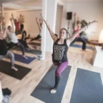 Eine Gruppe von Menschen macht Yoga-Übungen in einem hellen Raum, während eine Frau im Vordergrund in einer Pose verharrt.