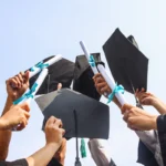 Mehrere Absolvent:innen halten ihre Diplomrollen und Hüte jubelnd in die Luft.