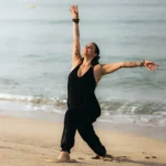 Eine Frau in schwarzer Kleidung im Yogaflow am Strand, während das Wasser sanft an den Sand spült.