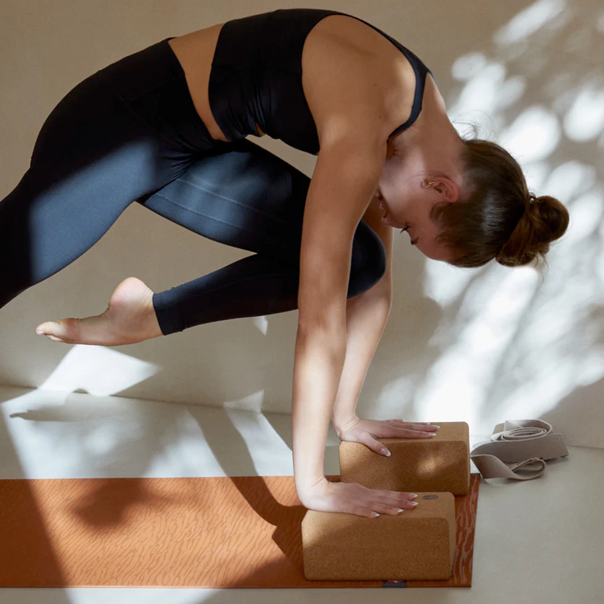 Frau in schwarzem Sporttop und blauen Leggings in einer nach vorne gebeugten Yoga-Pose, die Hände auf Korkblöcken abgestützt.
