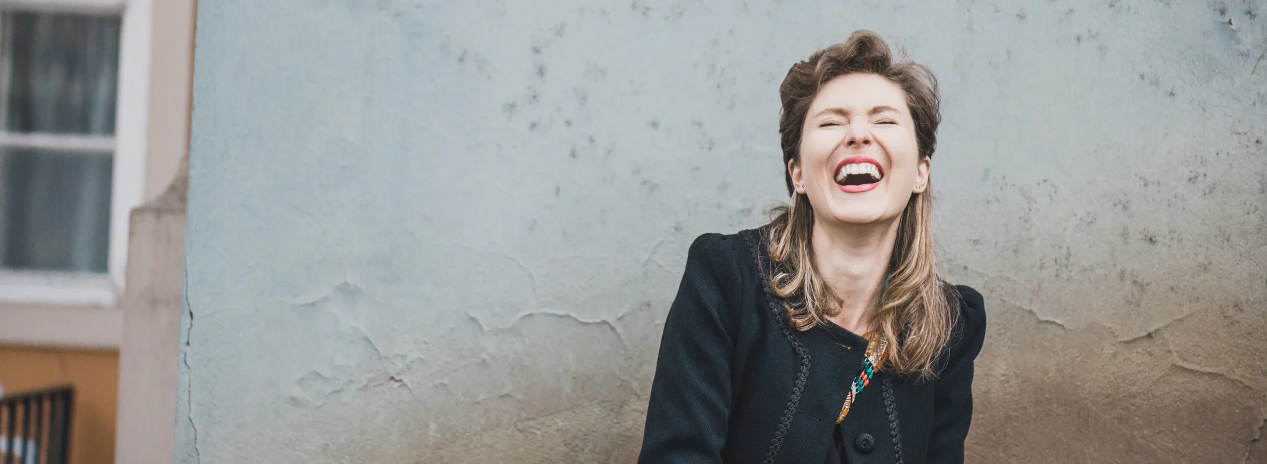 Eine lachende Frau in einem schwarzen Mantel steht vor einer verwitterten Wand und strahlt Freude aus.