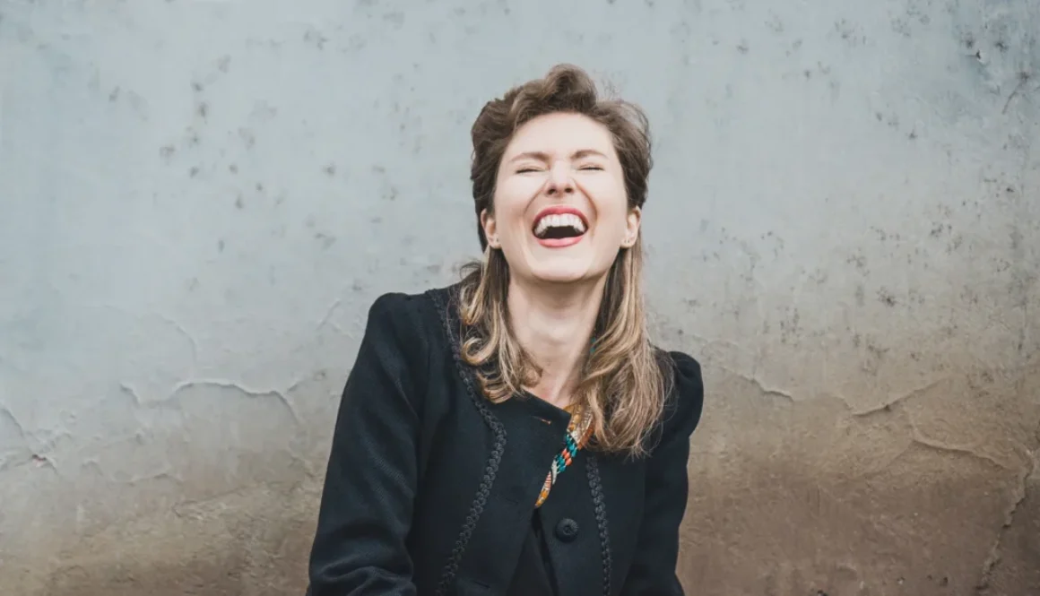 Eine fröhliche Frau lacht herzlich vor einer strukturierten Wand und strahlt pure Lebensfreude aus.