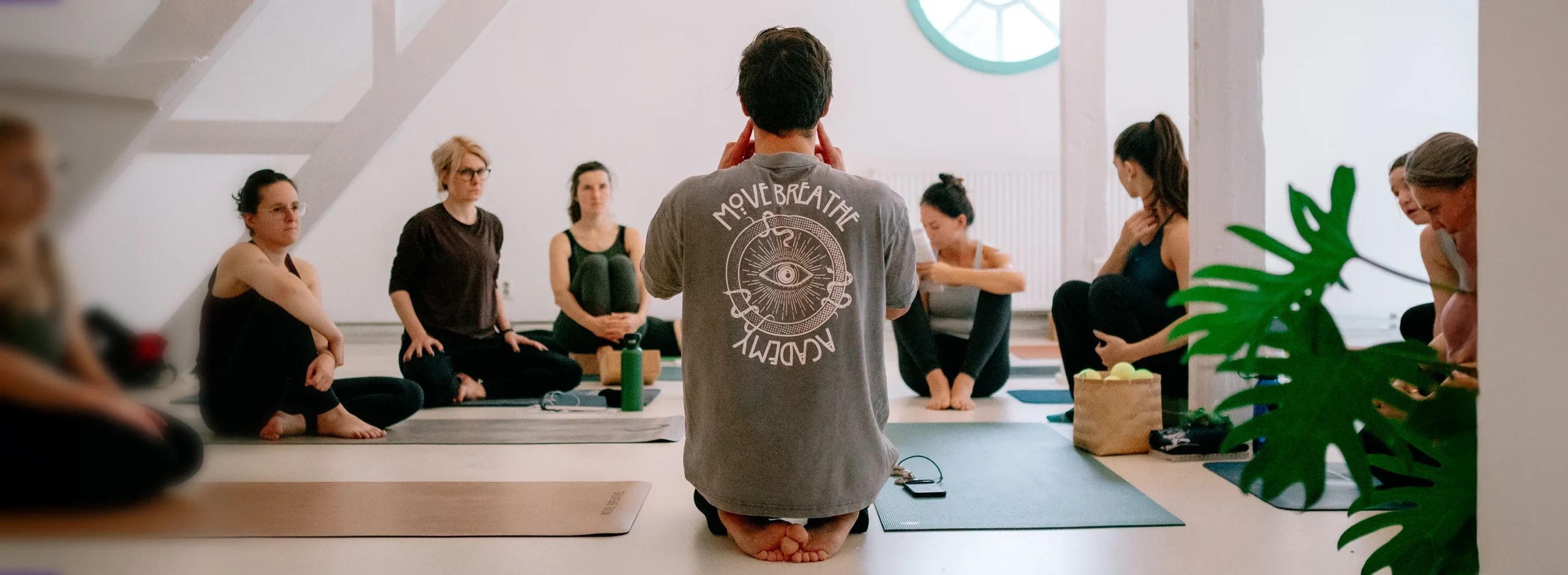 Eine Gruppe von Yogalehrer:innen sitzt aufmerksam im Kreis, während ein Lehrer Anweisungen gibt.