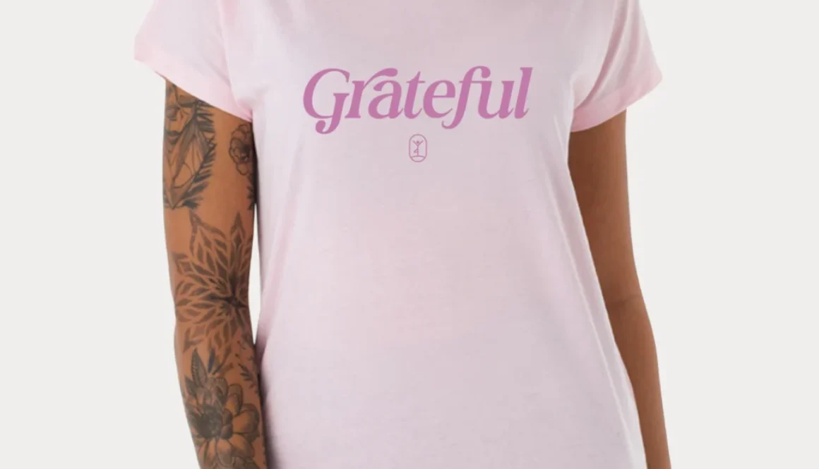 Eine Person trägt ein pinkes Shirt mit dem Aufdruck "Grateful" und zeigt stolz ihre tätowierten Arme.