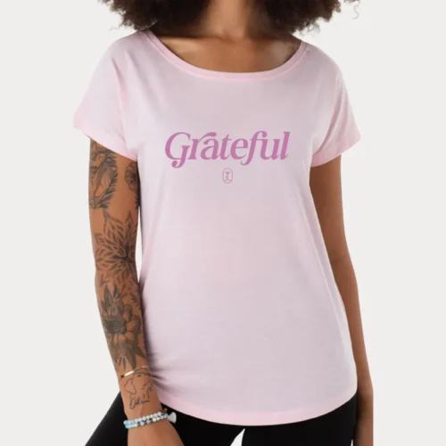 Eine Person trägt ein pinkes Shirt mit dem Aufdruck "Grateful" und zeigt stolz ihre tätowierten Arme.