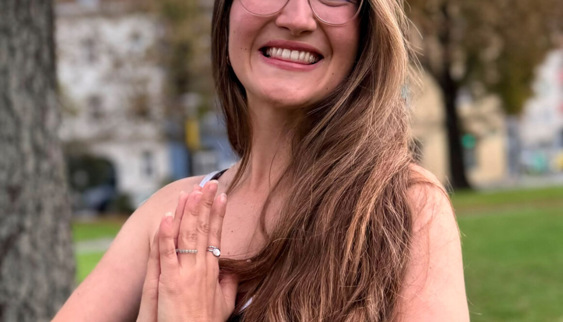 Michelle lächelt fröhlich, während sie im Freien in einer Yoga-Pose mit zusammengelegten Händen steht.