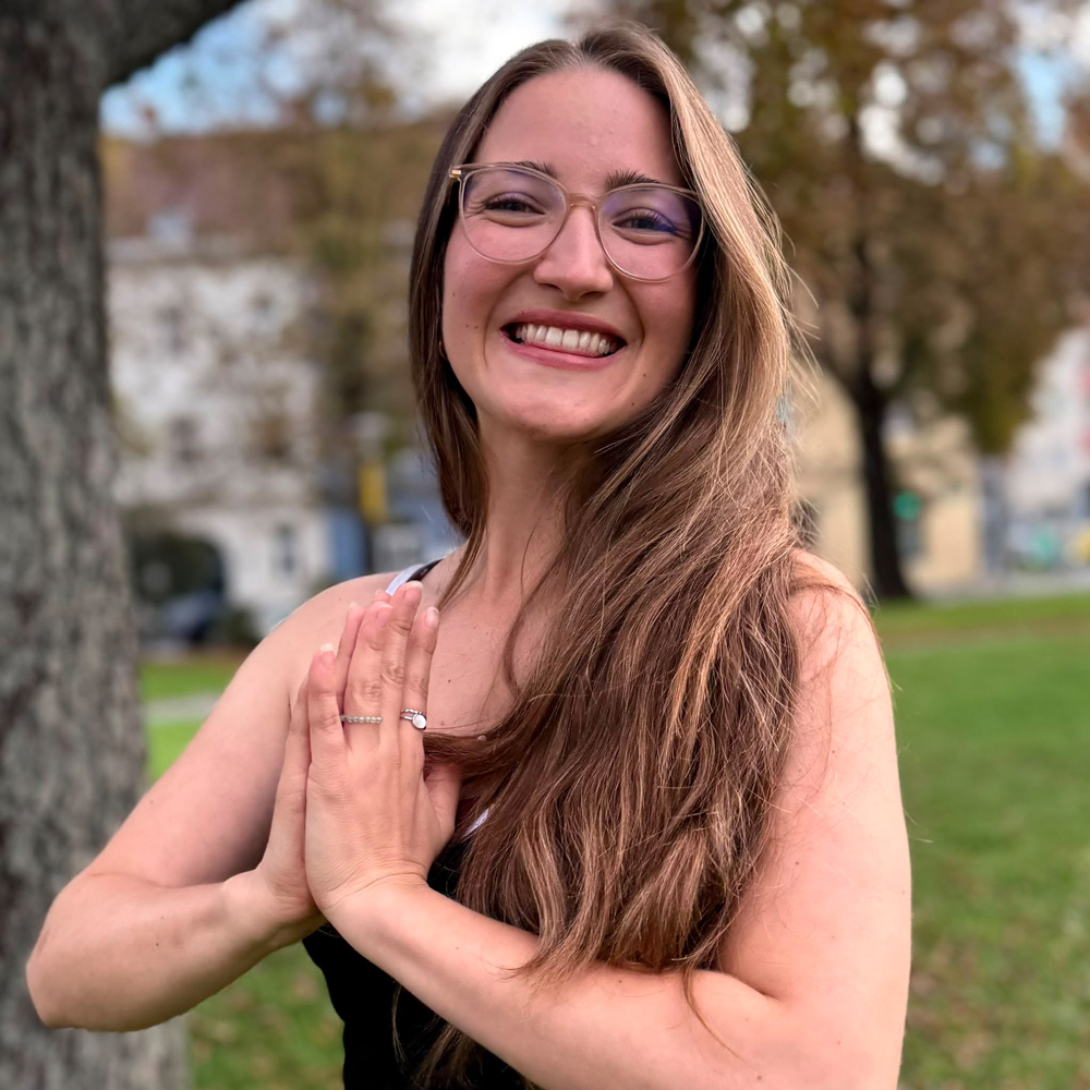 Michelle lächelt fröhlich, während sie im Freien in einer Yoga-Pose mit zusammengelegten Händen steht.