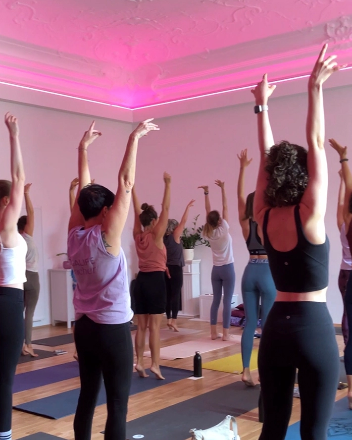 Die Gruppe praktiziert Yoga im YOGALIFE-Studio mit erhobenen Armen unter sanftem Licht.